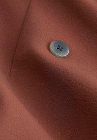 Un tissu couleur rouille avec une texture lisse présente un bouton rond, bleu clair, avec quatre trous, positionné près d'une couture.