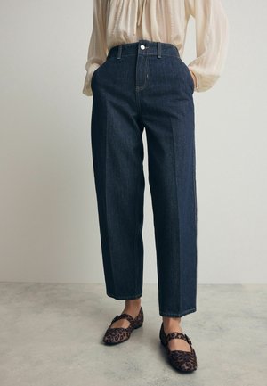 Osoba ubrana w ciemnoniebieskie, wysokogatunkowe jeansy z prostymi nogawkami, kremową bluzkę oraz baleriny Mary Jane w panterkę, stojąca na szarej podłodze.