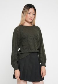 Ciemnozielony sweter z dzianiny z fakturowanym wzorem w zygzaki, z okrągłym dekoltem i długimi rękawami, zestawiony z plisowaną czarną spódnicą.