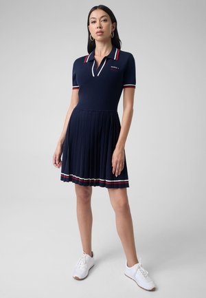 Mujer de pie que lleva un vestido tipo polo azul marino de manga corta con rayas rojas y blancas, falda plisada y zapatillas blancas.