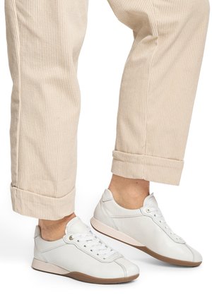 Feet wearing white leather lace-up sneakers with beige rubber soles, paired with rolled-up beige corduroy pants.