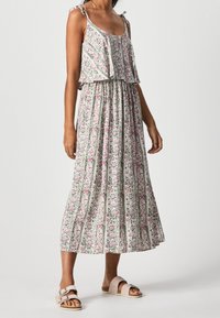 Mujer vistiendo un vestido midi sin mangas de estampado floral con detalles de volantes y sandalias rosa tipo slip-on con hebillas, de pie sobre un fondo liso.