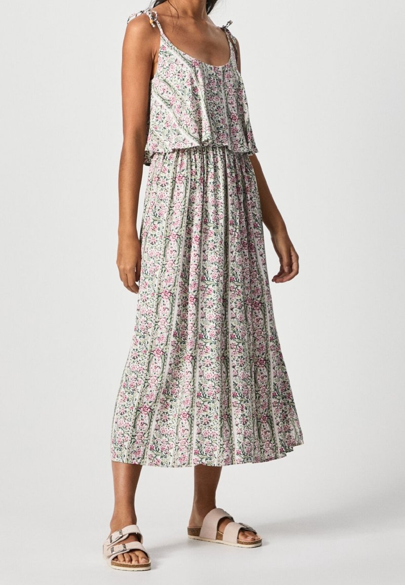 Mujer vistiendo un vestido midi sin mangas de estampado floral con detalles de volantes y sandalias rosa tipo slip-on con hebillas, de pie sobre un fondo liso.