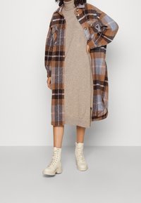 Manteau surdimensionné à carreaux marron et bleu, robe en maille beige en dessous, et bottes de combat crème. Comprend des boutons et deux poches poitrine.