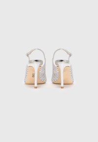 Et par sølvfargede høyhælte slingback-sko med glitrende stropper, tynne hæler og beige såler, sett fra baksiden på en hvit bakgrunn.