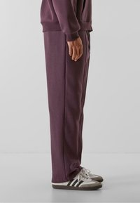 Lilla sweatpants med afslappet pasform, elastisk talje, sidelister og ribbede manchetter, parret med grå og hvide sneakers.