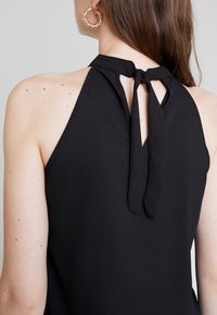 Haut noir à nouer derrière le cou avec fermeture par lien dans le dos. Tissu lisse avec un dos ouvert et une légère brillance. Boucle d'oreille en or visible.
