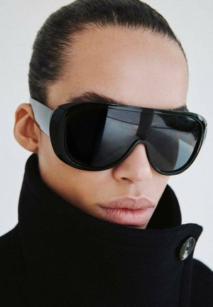 Gafas de sol negras de gran tamaño con lentes curvas y montura gruesa; detalles grises en las patillas; usadas con un abrigo negro de cuello alto.