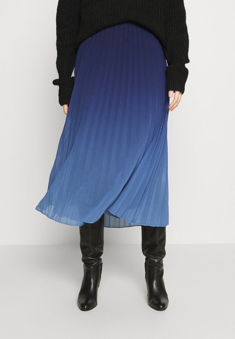 Femme portant une jupe midi plissée bleue, un pull noir et des bottes en cuir noires montantes, debout devant un fond uni.