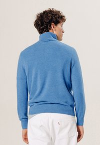 Helles blauer Rollkragenpullover aus weichem Material, figurbetontes Design, lange Ärmel sowie gerippte Bündchen und Saum. Getragen mit weißen Hosen.