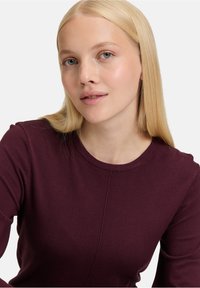 Burgunderfarbener Pullover mit Rundhalsausschnitt, figurbetonten Ärmeln und glatter Textur; weist subtile Steppdetails auf.