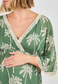 Grünes Kleid mit weißem Palmbaumdruck, V-Ausschnitt, Spitzenverzierung an den Ärmeln, leichtes, fließendes Material, geraffte Taillenpartie.