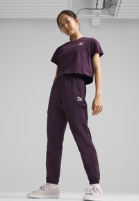 Violet viršus ir derinamos kelnės, pagamintos iš lengvos medžiagos, su mažai baltu logotipu ant krūtinės ir kelnių. Baltos sportbačiai puikiai užbaigia įvaizdį.
