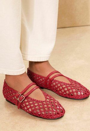 Feet wearing red woven flat shoes with double straps and buckles, paired with off-white pants.