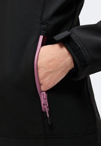 Main partiellement dans la poche de la veste noire avec une fermeture éclair rose visible et un poignet de manche ajustable avec une bande Velcro noire.