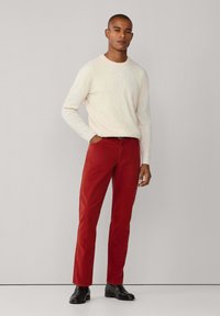 Hombre de pie vistiendo un suéter blanco de punto trenzado, pantalones rojos, cinturón negro y zapatos de vestir negros, frente a un fondo claro y sencillo.