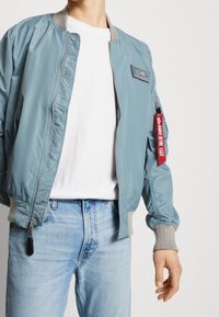 Light blue bomber jacket with a zip front, gray ribbed collar and cuffs, side pockets, and a red patch on the sleeve for added detail.