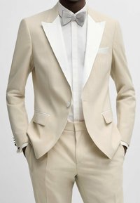 Uomo in smoking beige con rever bianchi, camicia bianca e cravatta a farfalla in tessuto argentato, mani nelle tasche.