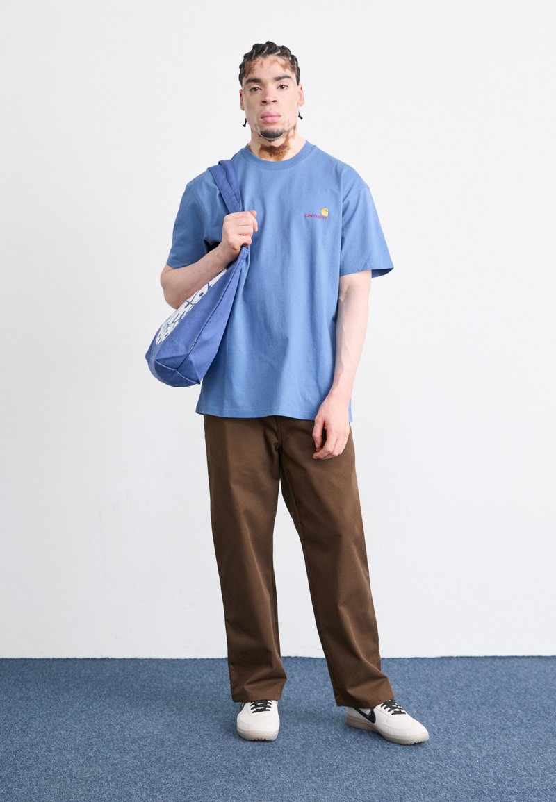 T-shirt di cotone blu con logo, pantaloni marroni e sneaker bianche. Presenta una borsa blu con design grafico, indossata a spalla.