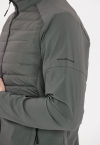 Grå jacka med en vadderad framdel, släta ärmar och en dragkedja vid kragen. Ordet "ENDURANCE" är tryckt på vänster ärm.