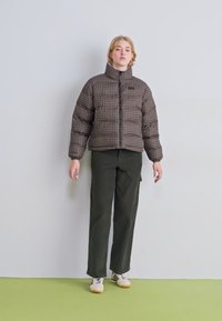 Hnědá kostkovaná péřová bunda, zapínání na zip, vysoký límec; v kombinaci s tmavozelenými širokými kalhotami a světle hnědými teniskami, na světlém pozadí.