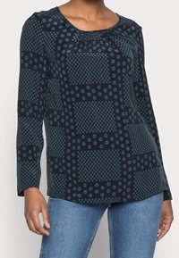 Blusa navy realizzata in tessuto leggero, con motivo circolare e quadrato in teal. Maniche lunghe con scollatura rotonda e dettaglio arricciato.