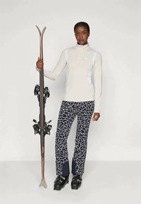 Γυναίκα με λευκή μπλούζα μακρυμάνικη και patterned παντελόνι σκι στέκεται κρατώντας ένα ζευγάρι σκι, φορώντας μαύρες μπότες σκι, σε απλή φόντο.