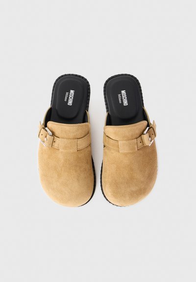 Tan mockasiner i mocka med justerbara sidspännen och svarta innersulor märkta "Moschino Milano" på en ljus bakgrund.