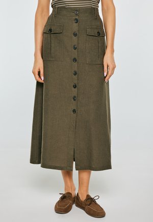 Kobieta w oliwkowozielonej spódnicy midi zapinanej na guziki z przodu, z przednimi kieszeniami na klapę, oraz w brązowych mokasynach na sznurowadła.