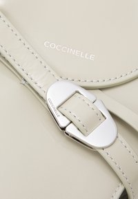 Skórzana torba marki Coccinelle w jasnobeżowym kolorze z białym przeszyciem, wyposażona w srebrną klamrę oraz logo wytłoczone na powierzchni.