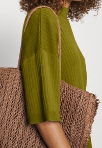 Zelený žebrovaný svetr s tříčtvrtečními rukávy, doplněný tkanou hnědou taškou s třepenými okraji.