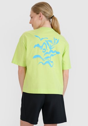 Kobieta z blond kucykiem ubrana w limonkową koszulkę z abstrakcyjnym niebieskim wzorem natury na plecach i czarne szorty, zwrócona tyłem.