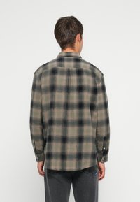 Camisa de franela en un patrón de cuadros en beige apagado, negro y gris. Presenta un collar, mangas largas y puños con botones. La parte posterior tiene un detalle de yugo.