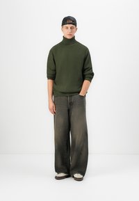 Junger Mann mit einer schwarzen, verkehrtherum getragenen Mütze, einem grünen Rollkragenpullover, weiten schwarzen Jeans und weißen Sneakers, der vor einem weißen Hintergrund steht.