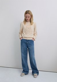 Bluză bej cu un design cu craniu și oase încrucișate, asortată cu blugi largi bleu deschis, decorați cu modele brodate. Adidași strălucitori.