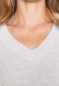 Pull en tricot gris clair avec un design en V. La texture est lâche et aérée, avec un col en rib et un tissage subtilement ouvert.