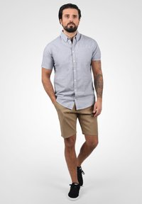 Camisa de botones de manga corta azul claro, hecha de tela tejida, combinada con pantalones cortos beige. Zapatillas negras realzan el aspecto informal.