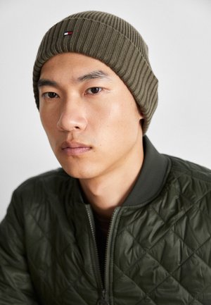 Olivgrüner, gerippt gestrickter Beanie mit kleinem Logo, getragen von einer Person in einer gesteppten grünen Bomberjacke mit Rautenmuster und Reißverschluss.