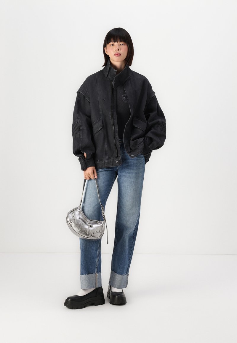 Jeune femme portant une grande veste noire, un jean bleu retroussé, des chaussures noires épaisses, tenant un petit sac à main en argent, se tenant contre un fond blanc.