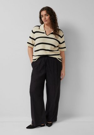 Vrouw met krullend haar die een korte mouwen crèmekleurige trui met zwarte strepen draagt, zwarte broek met wijde pijpen en zwarte puntige schoenen.