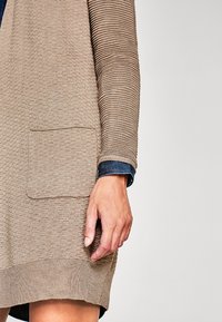 Beige cardigan med teksturerede striber, som har en forlommer og en ribstrikket kant. Blå denim underlag synlig ved manchetten.