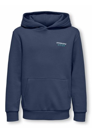 Garçon aux cheveux afro portant un sweat à capuche bleu marine, un pantalon de survêtement gris et des baskets blanches, debout les mains sur les hanches devant un fond uni.