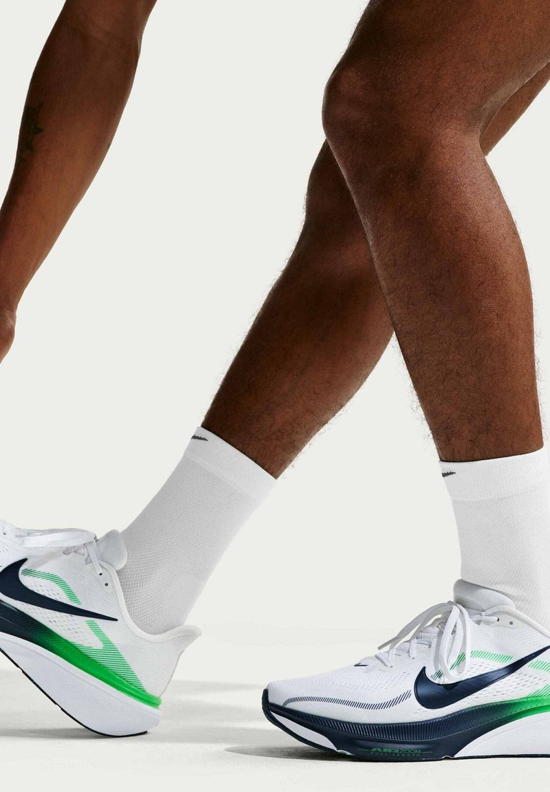 Πόδια με λευκά αθλητικά παπούτσια Nike με πράσινες και μαύρες λεπτομέρειες, συνδυασμένα με λευκές κάλτσες, βήμα προς τα εμπρός σε φωτεινό φόντο.