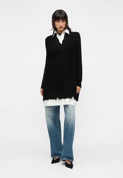 Μαύρο πλεκτό πουλόβερ με V-λαιμόκοψη και ξεφτισμένο τελείωμα, φορεμένο πάνω από λευκό πουκάμισο. Συνδυάζεται με ανοιχτό μπλε ξεθωριασμένο τζιν και μαύρα μυτερά μπαλαρίνες.