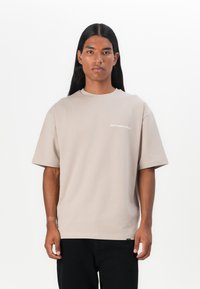 Beige bomulds T-shirt med korte ærmer, rund hals og hvid tekst "spild ikke kultur" på forsiden. Båret med sorte bukser.