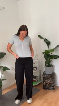 Top henley gri cu mânecă scurtă și ribbed, cu trei nasturi, purtat cu pantaloni negri lejeri și adidași albi, stând pe un podea din lemn.