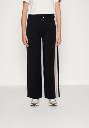 Osoba ubrana w czarne spodnie szerokie z różowo-białymi paskami po bokach i srebrne trampki, stojąca na jednolitym tle.