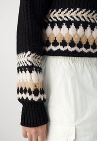 Czarny dziergany sweter z wzorzystym mankietem ozdobionym beżowymi, białymi i czarnymi rombami. Materiał jest gruby i prążkowany, zestawiony z jasnymi spodniami.