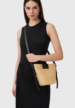 Woven straw bucket bag with a flat black leather accent and adjustable black strap, held by a person in a fitted black dress.