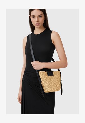 Vevet halmfutteral med en flat svart lærdetalj og justerbar svart stropp, holdt av en person i en tettsittende svart kjole.